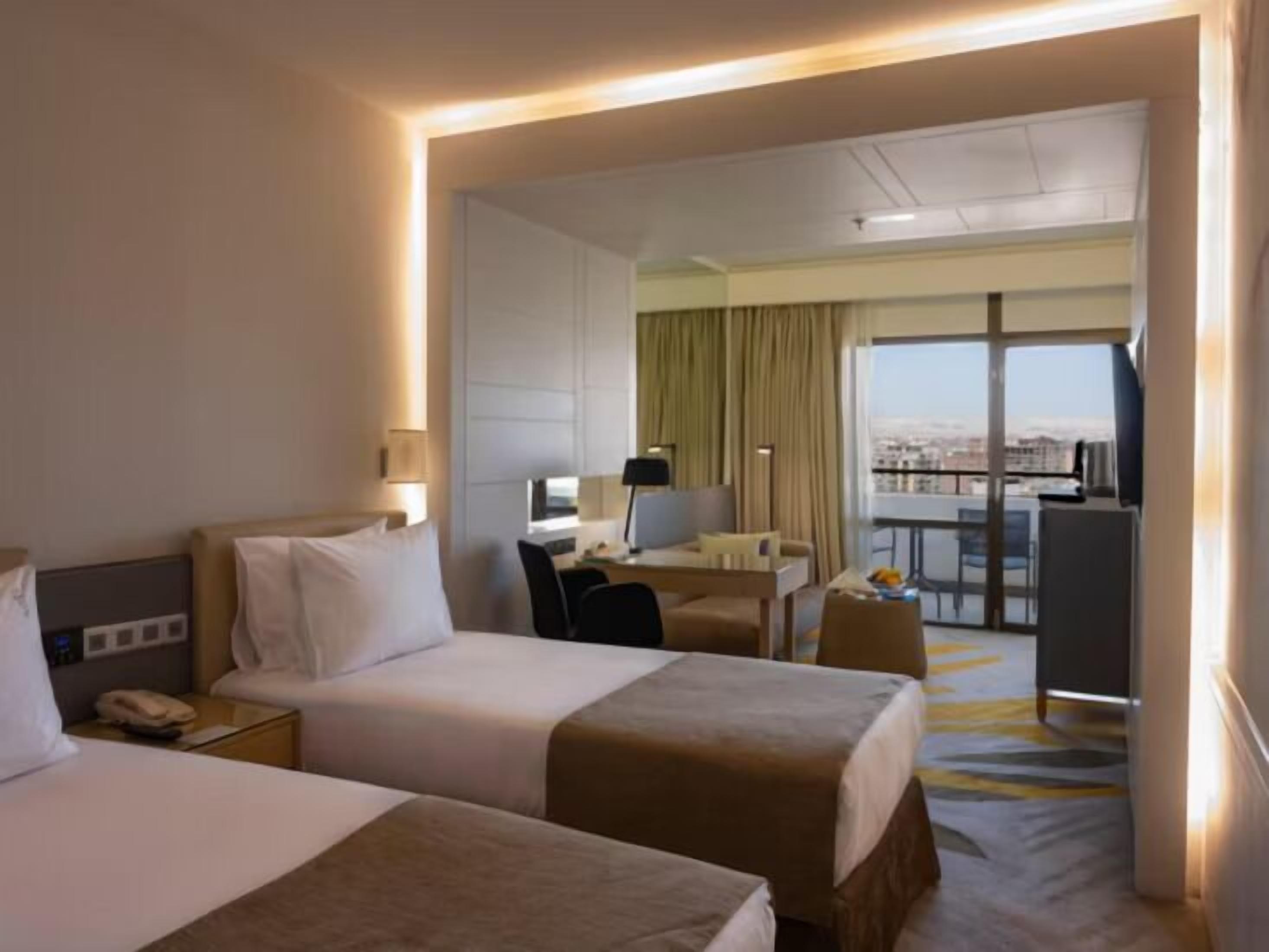 Ein Hotelzimmer mit zwei Betten, einem Schreibtisch, einem Stuhl, einem Telefon und Blick auf die Stadt durch große Fenster.