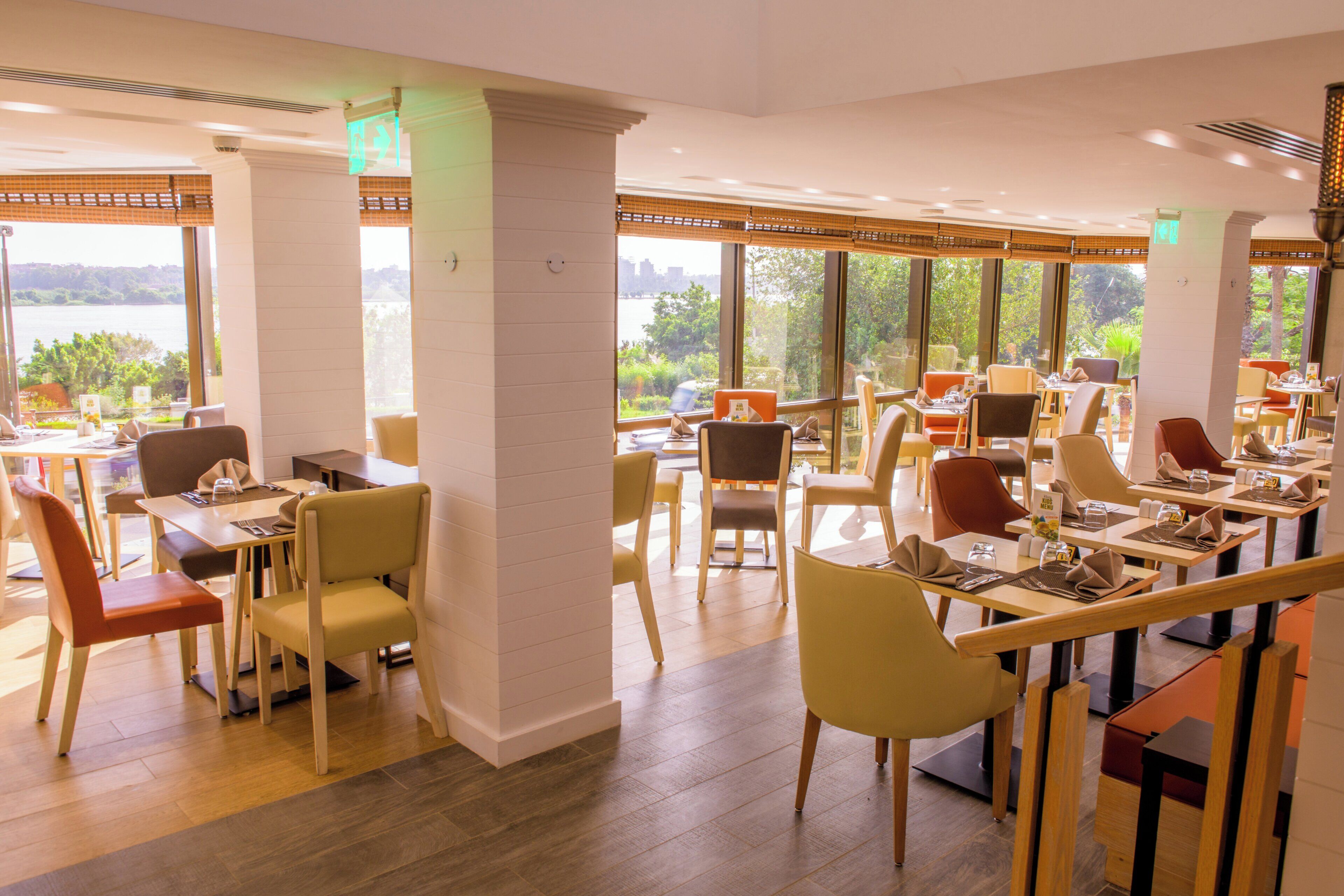 A restaurant with multiple tables and chairs, large windows with a view of greenery, and a modern interior design.