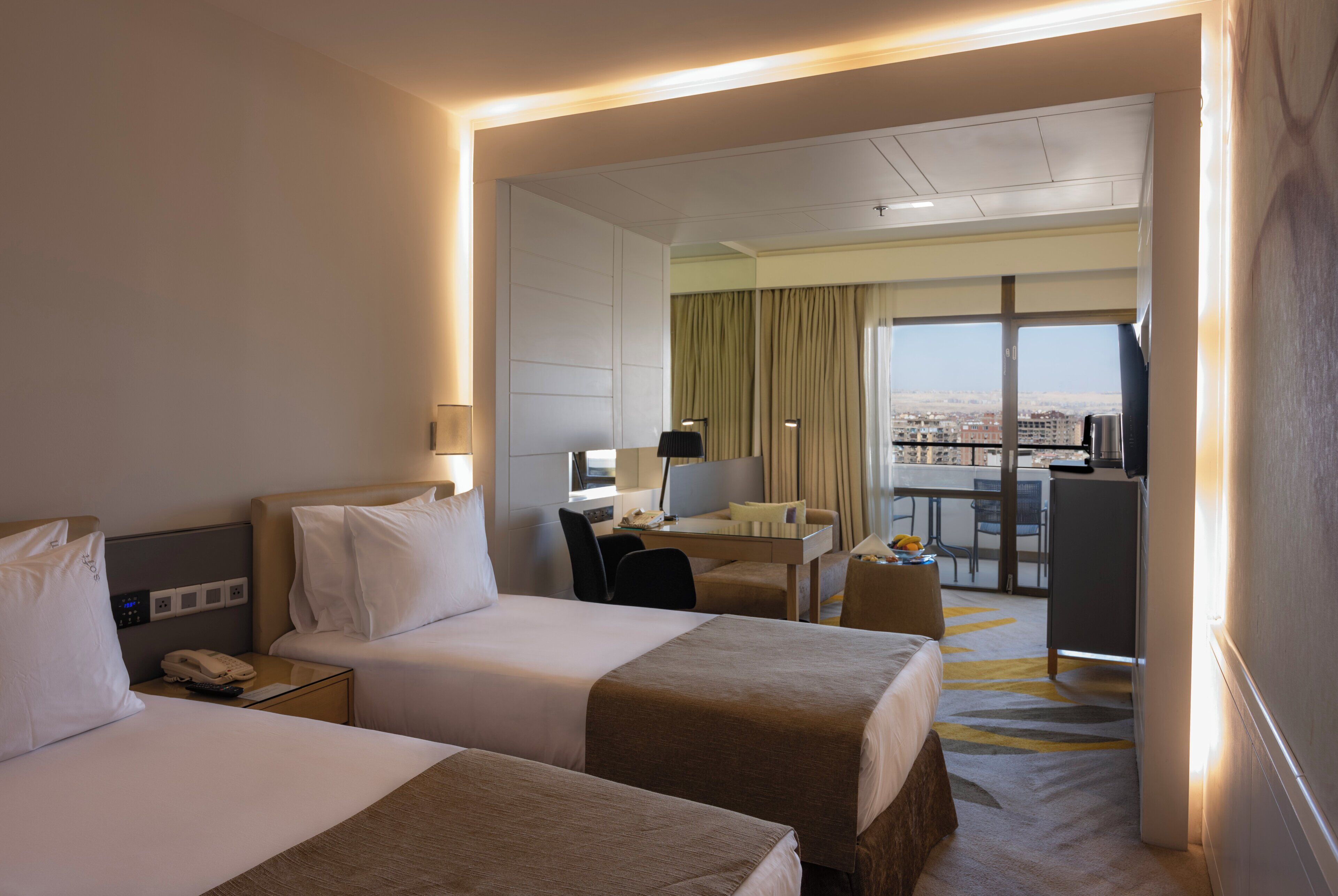 Habitación de hotel con dos camas, un escritorio, una silla, una mesita y vistas a la ciudad a través de grandes ventanales.