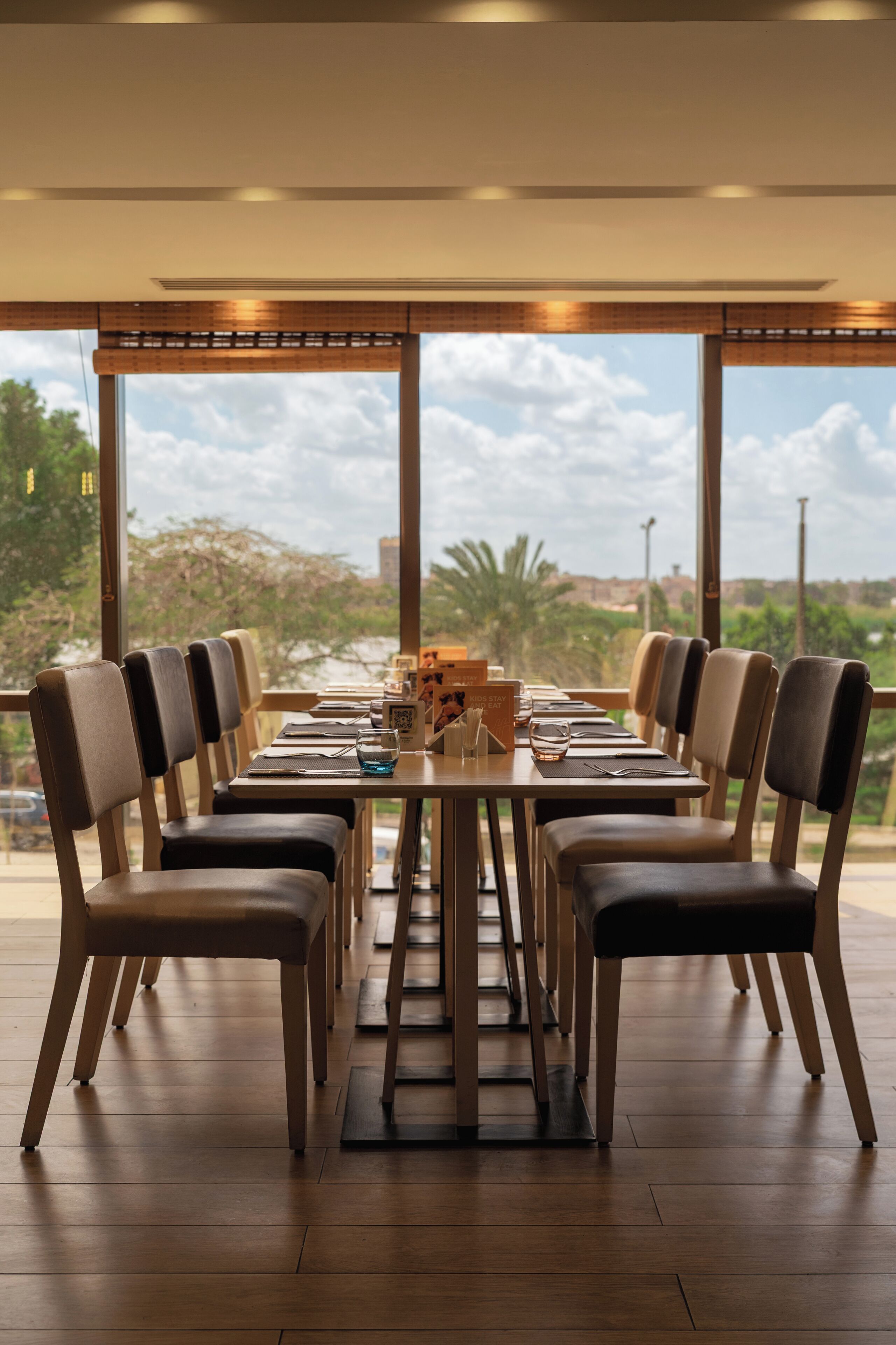 A dining area with a long table and chairs, large windows with a view of greenery and buildings, and a ceiling with recessed lighting.