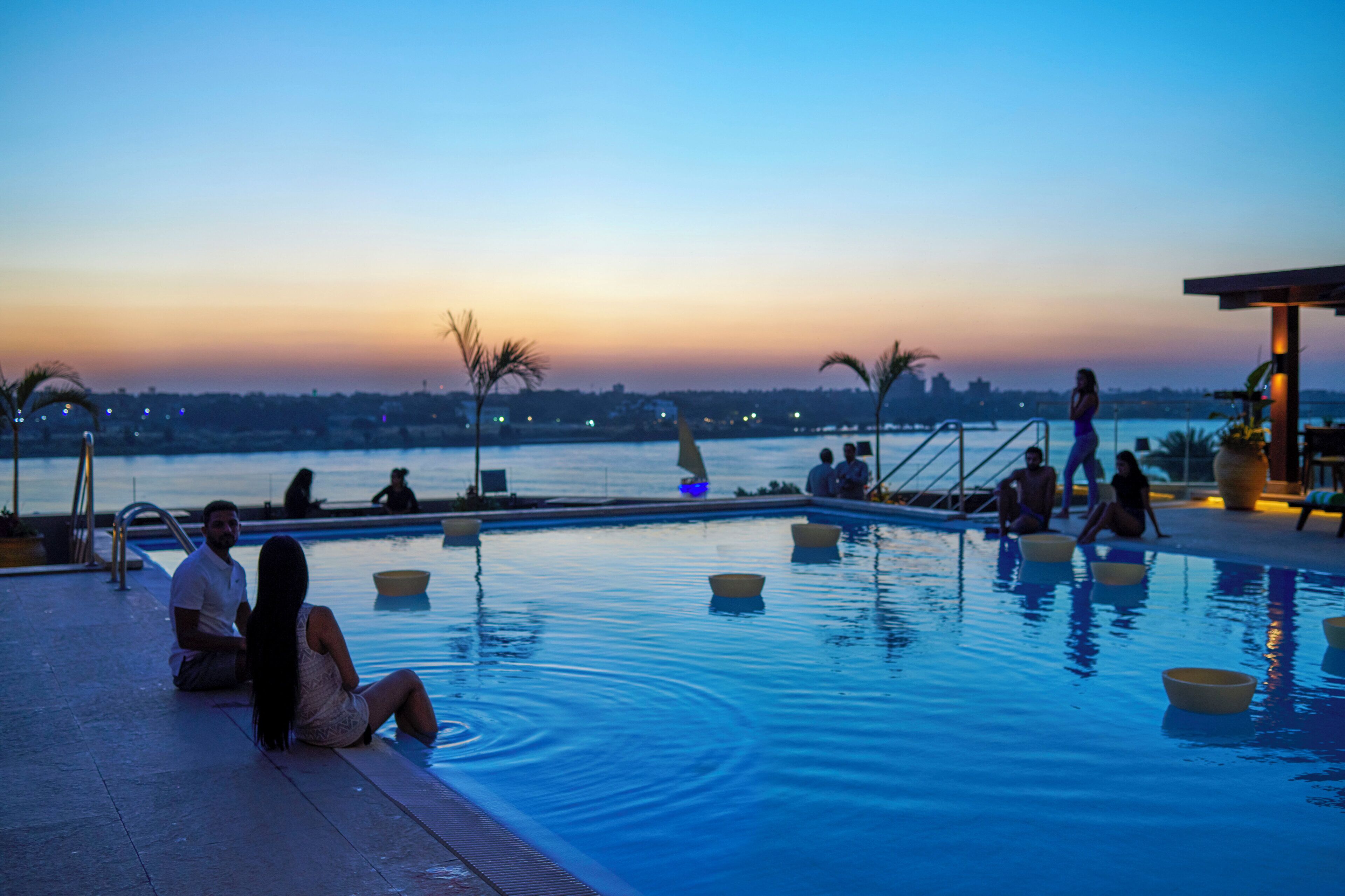 Una piscina con gente relajándose, un atardecer de fondo y vistas a la ciudad.