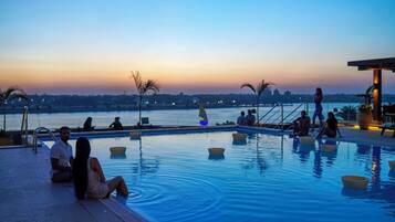 Una piscina con gente relajándose, un atardecer de fondo y vistas a la ciudad.