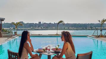 Two individuals seated at a table by a poolside with a cityscape in the background.