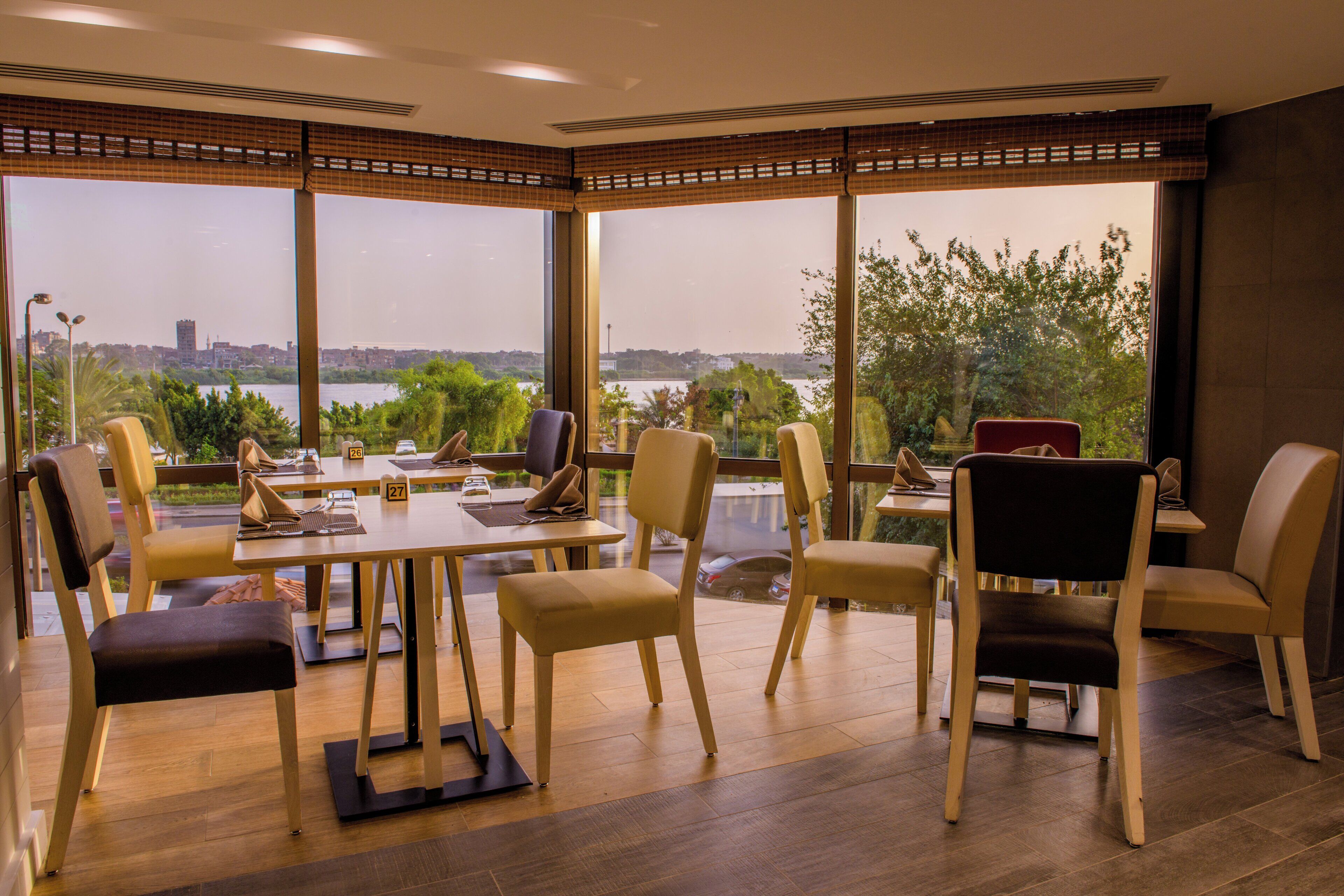 A dining area with tables and chairs, large windows with a view of a cityscape and greenery.