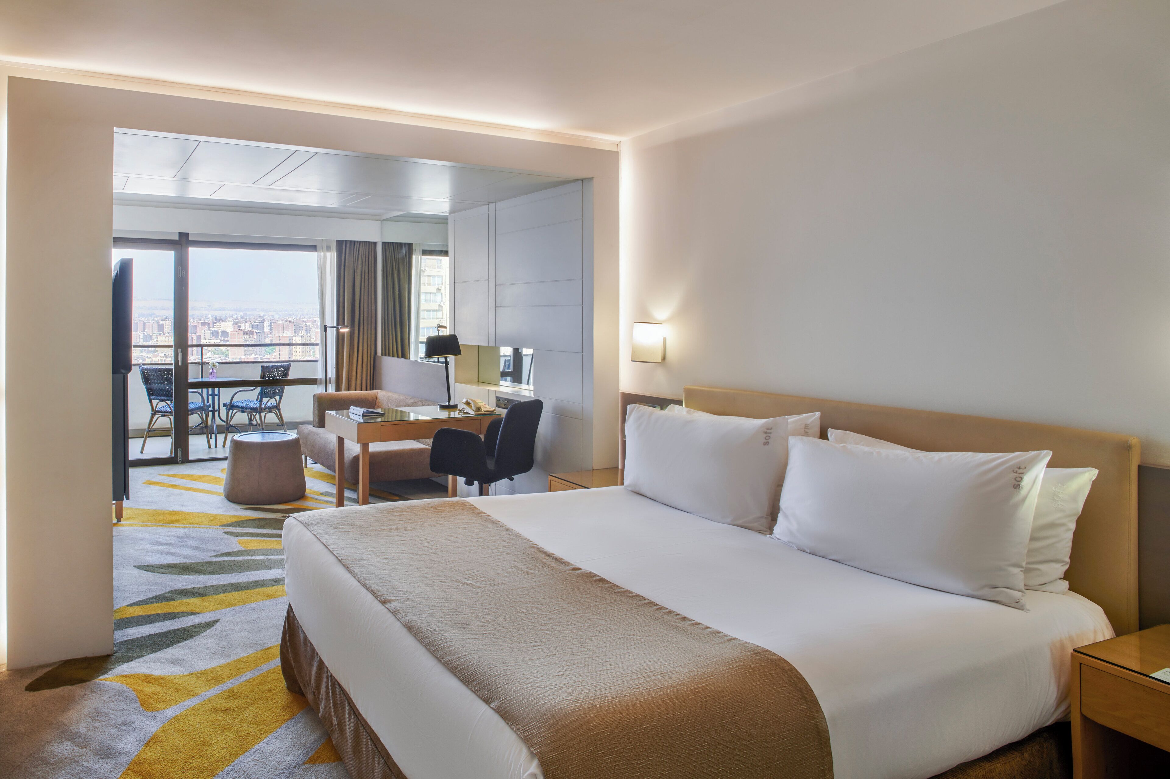 A hotel room with a large bed, a desk with a chair, a small round table, and a balcony with a view.