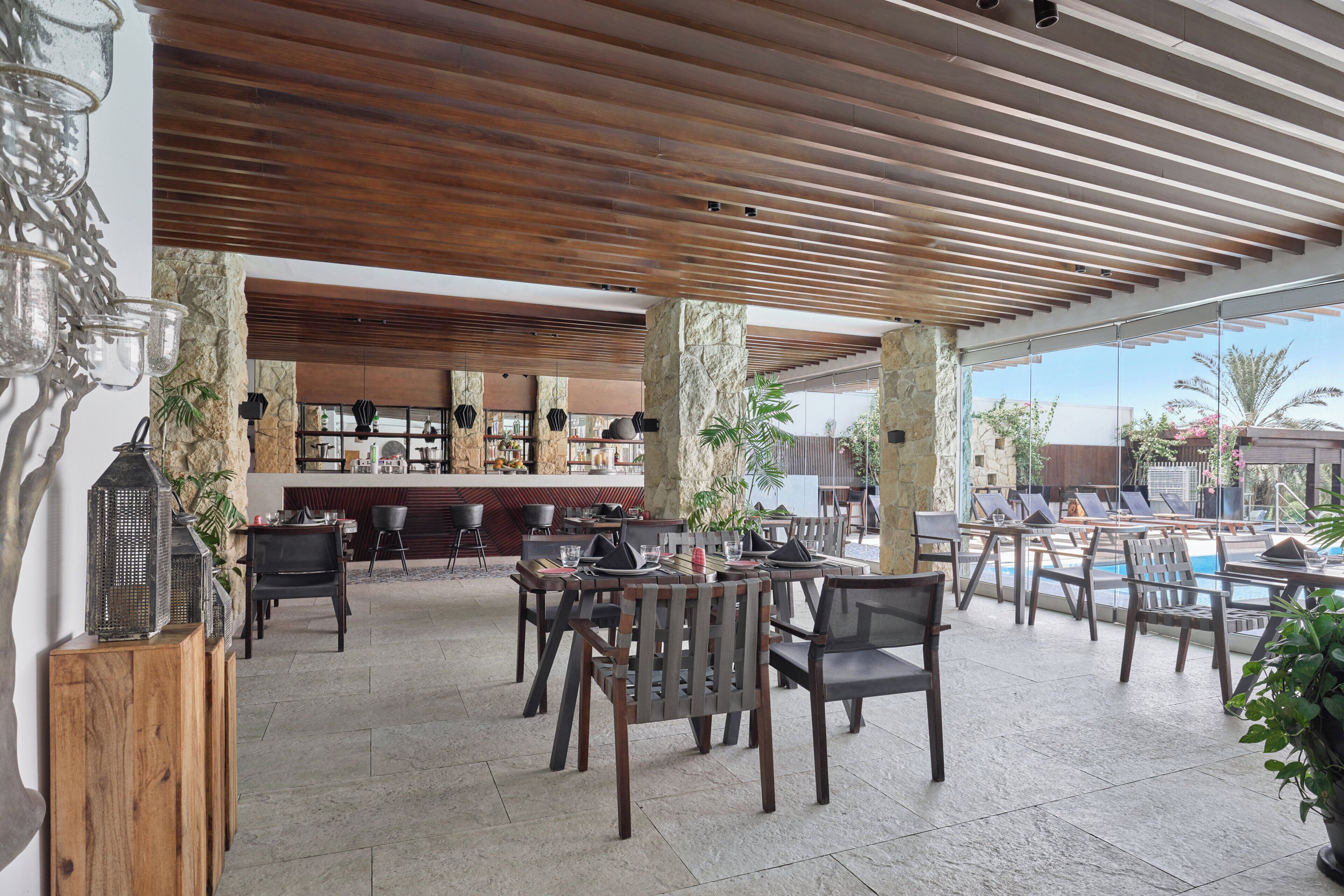 Een overdekt buitengebied met tafels, stoelen, een zwembad en een bar met een stenen muur en planten.