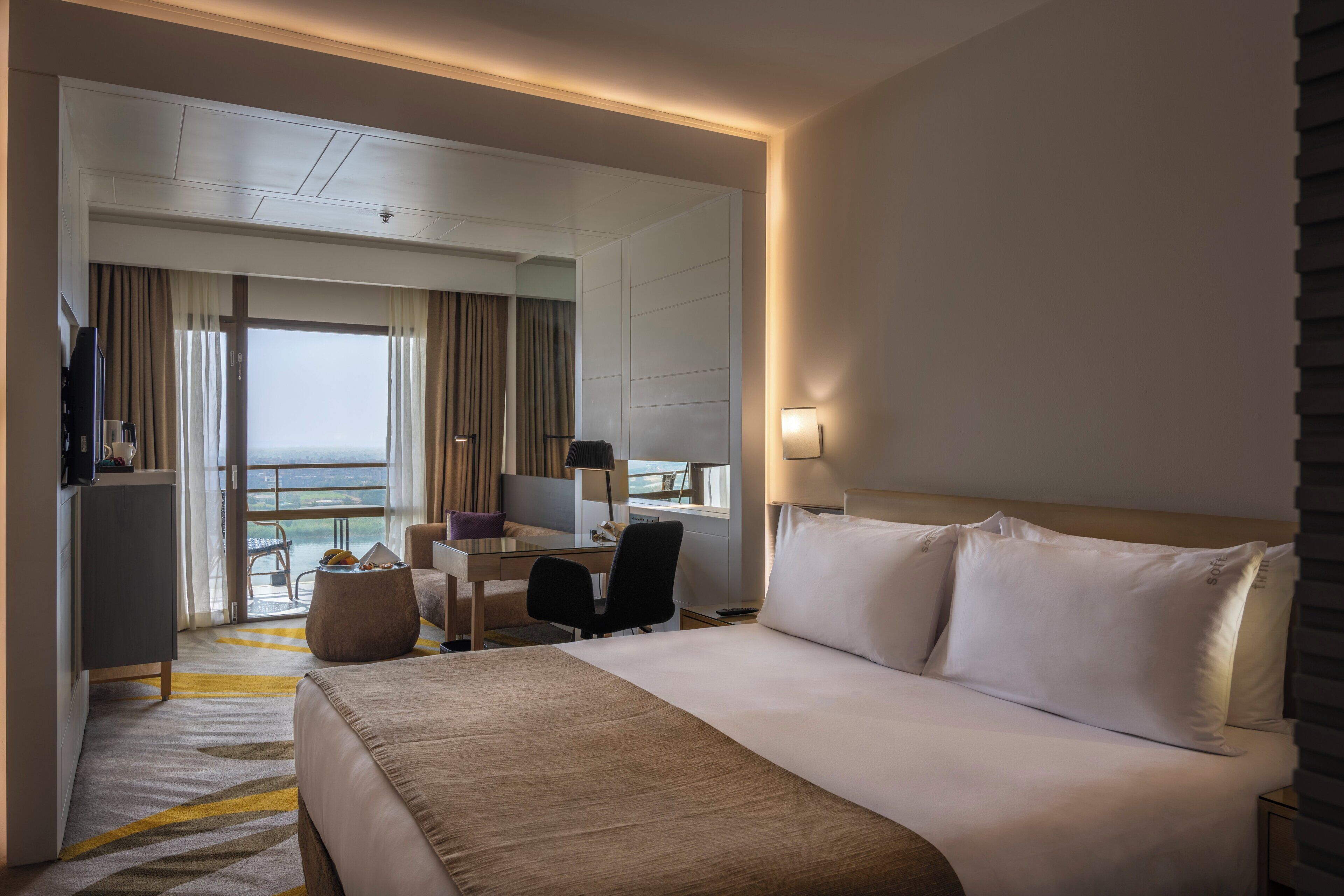 A hotel room with a large bed, a desk, a chair, and a view of the sea through a sliding door.