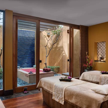 A spa room with two massage tables, a wooden floor, a small garden view, and a sliding glass door leading outside.