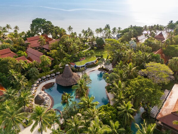 A resort with a central swimming pool, palm trees, and thatched-roof buildings.