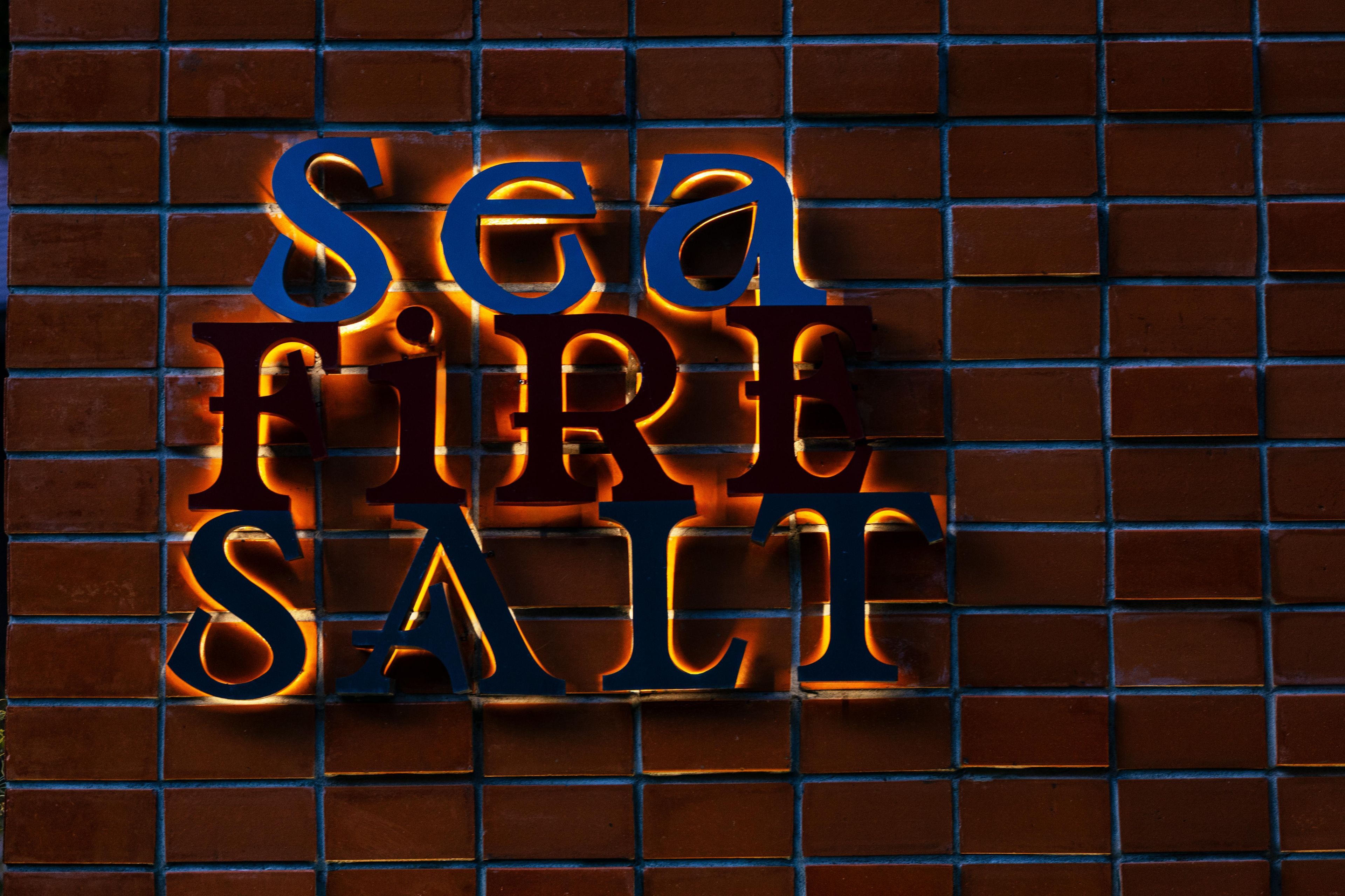 Een bord met de woorden 'SEA FIRE SALT' in een gestileerd lettertype, voor een achtergrond van een bakstenen muur.