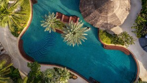 Vista aérea de un complejo turístico con una amplia piscina central, rodeado de palmeras y áreas de descanso con sombrillas.