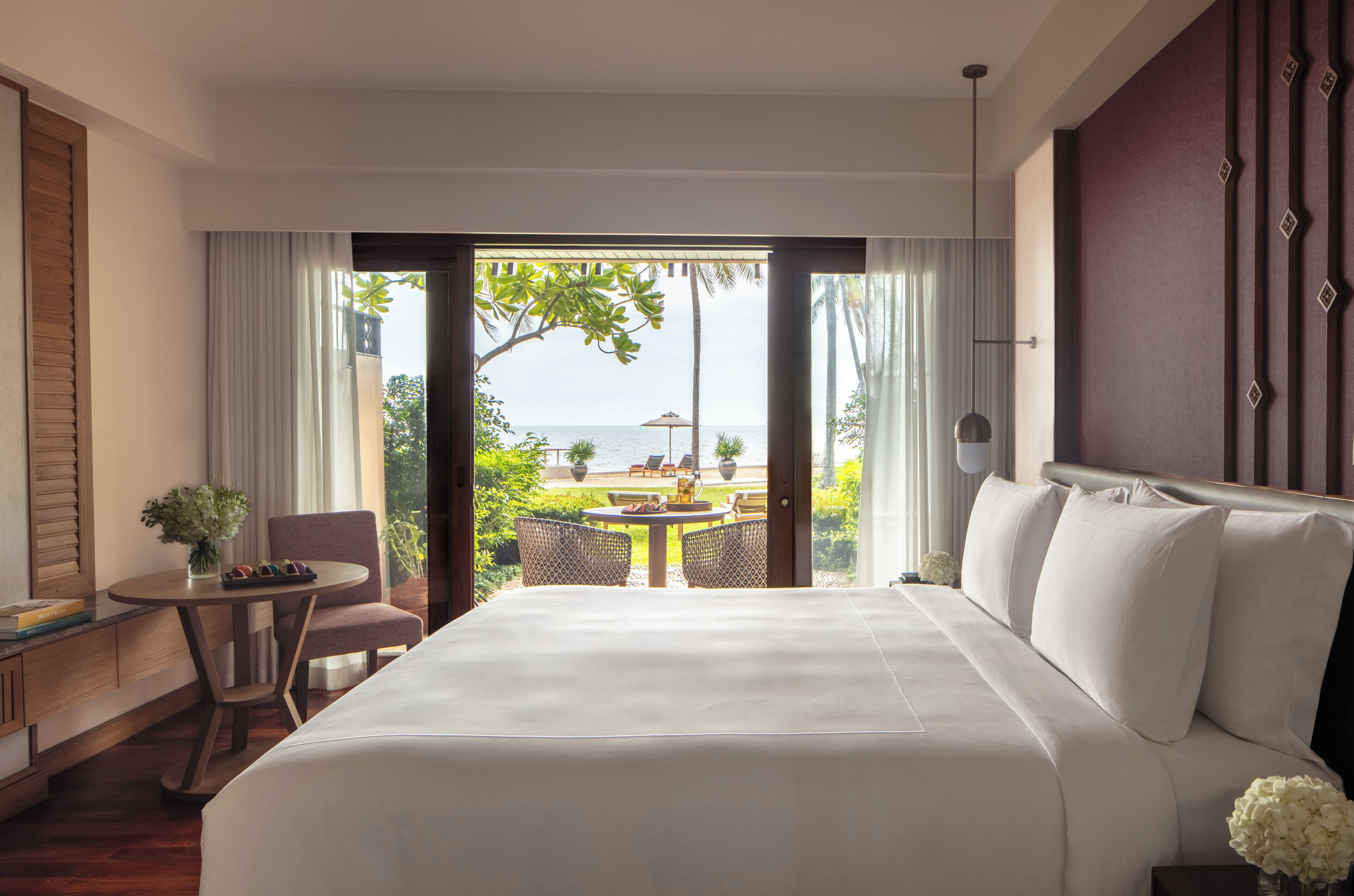 A hotel room with a large bed, a small table with chairs, a view of the sea, and a balcony with outdoor furniture.