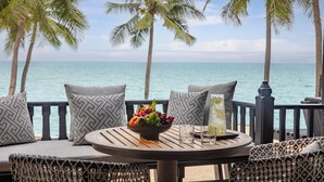 A round wooden table with a bowl of fruit, two glasses, and a bottle on a balcony overlooking the sea with palm trees.