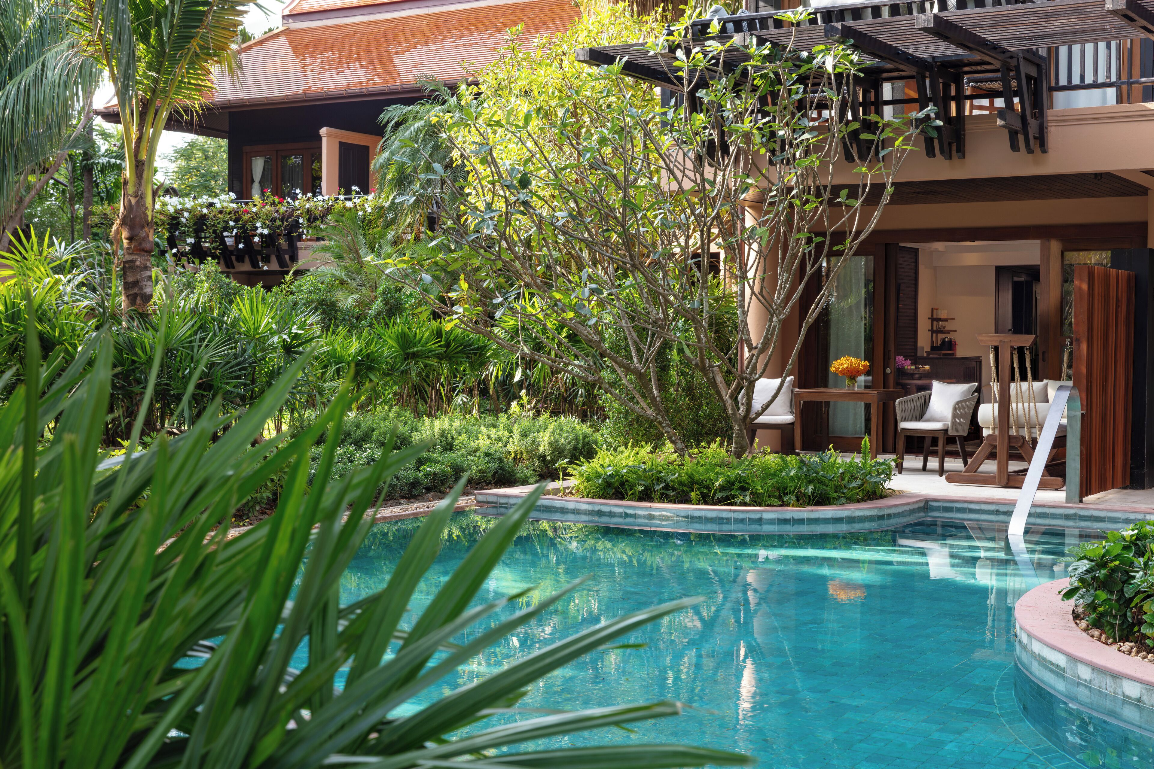 A pool surrounded by lush greenery, a wooden deck, and a building with a tiled roof.