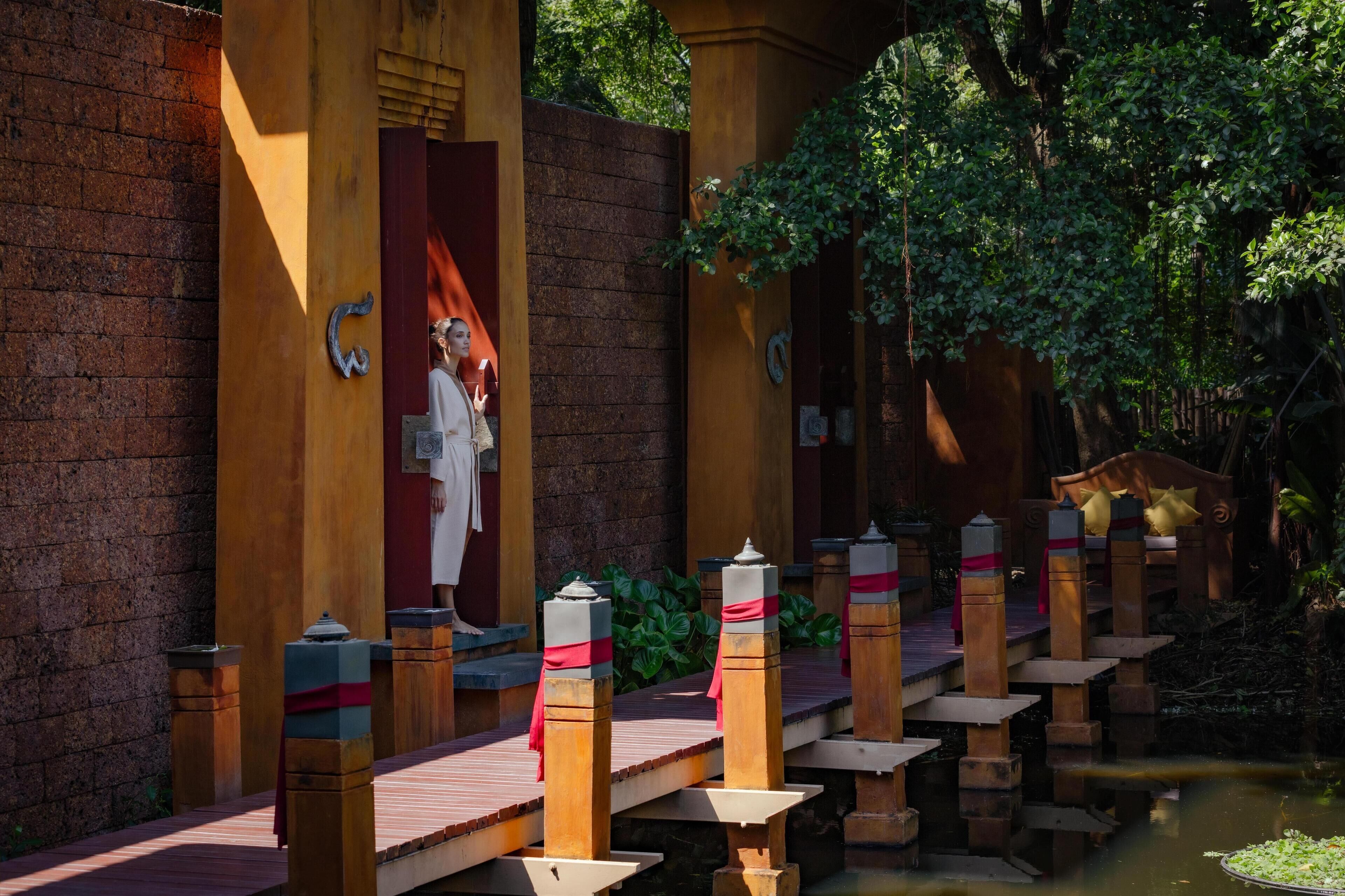 A traditional wooden bridge with a red and white railing, leading to a building with a large red door and a statue.
