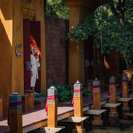 A traditional wooden bridge with a red and white railing, leading to a building with a large red door and a statue.