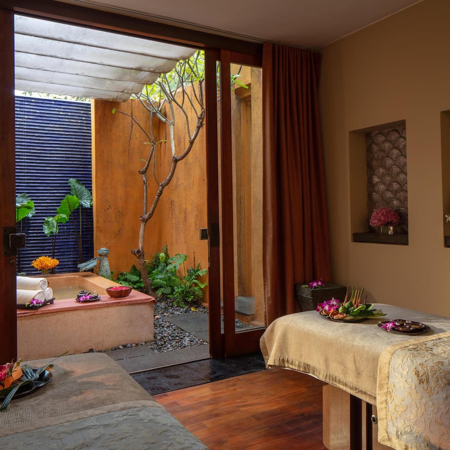 A spa room with a massage table, a small sitting area with a bench and cushions, a window with a view of a garden, and a wall with a decorative panel.