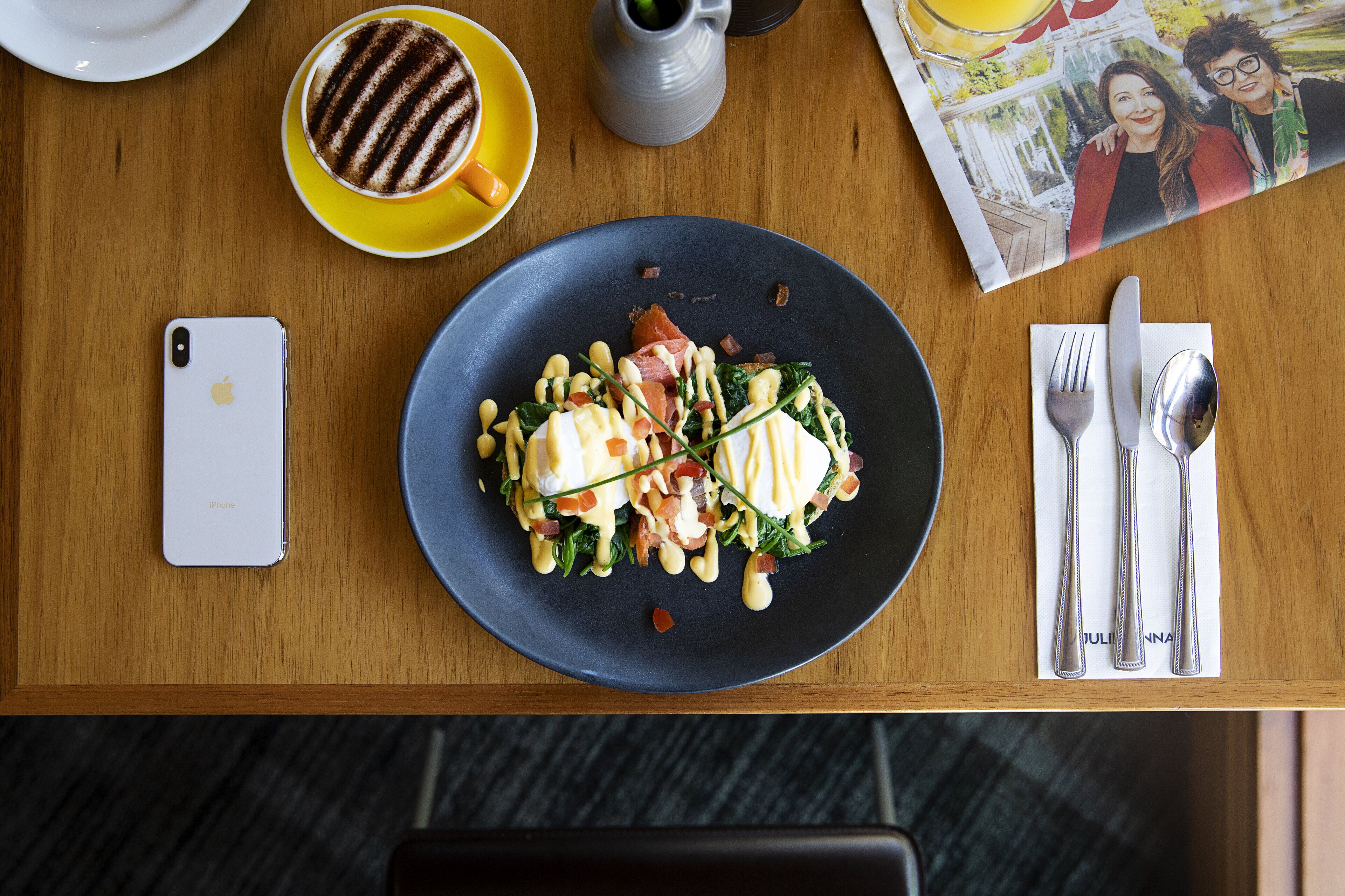 A dining table with a plate of food, cutlery, a smartphone, a coffee cup, and a magazine.