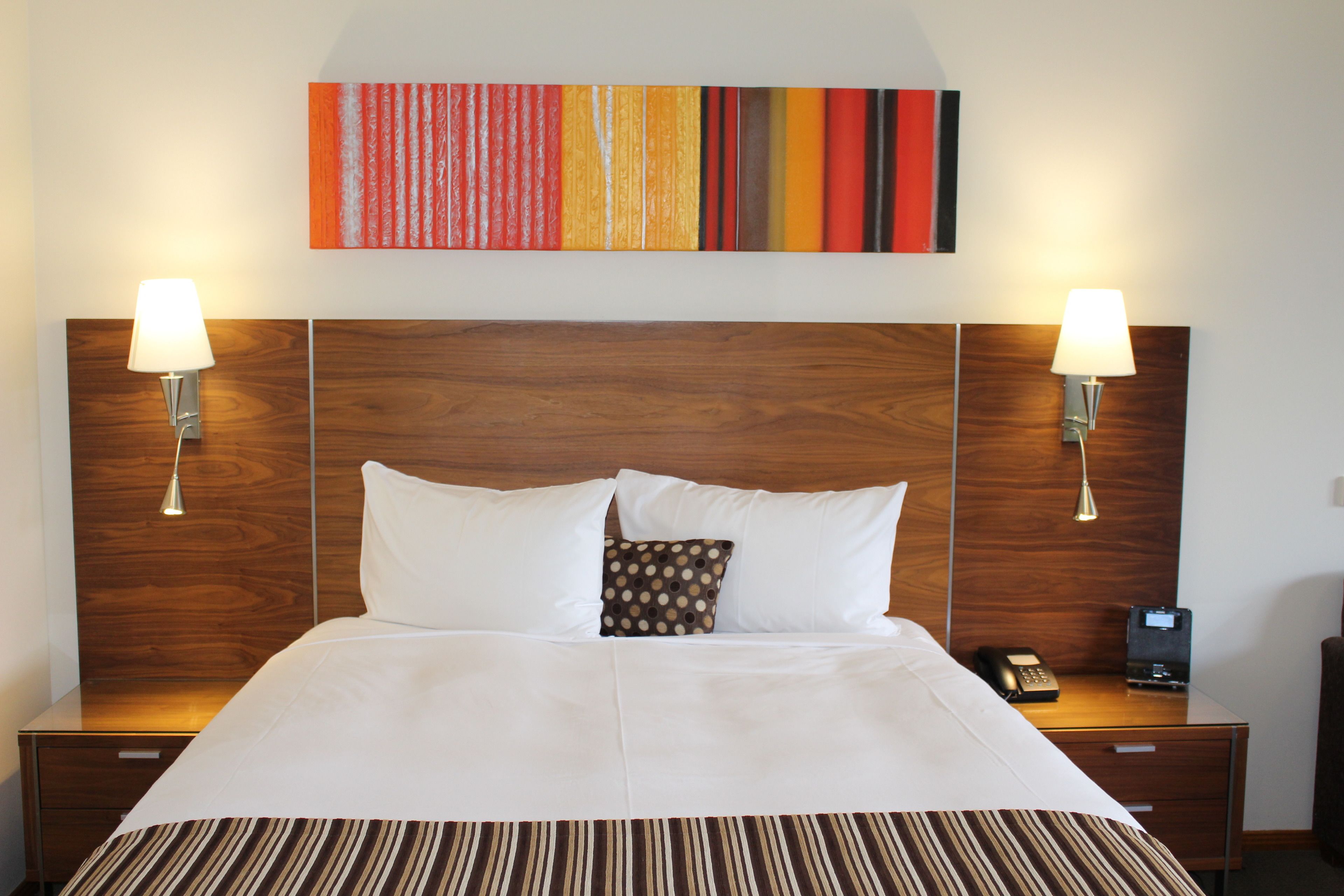 Ein Hotelzimmer mit einem Holzkopfteil, zwei Nachttischlampen, einem Bett mit weißen Bettwäsche, einem Streifen-Teppich, einem kleinen Zierkissen, einem Telefon und einer Kaffeemaschine auf dem Nachttisch.