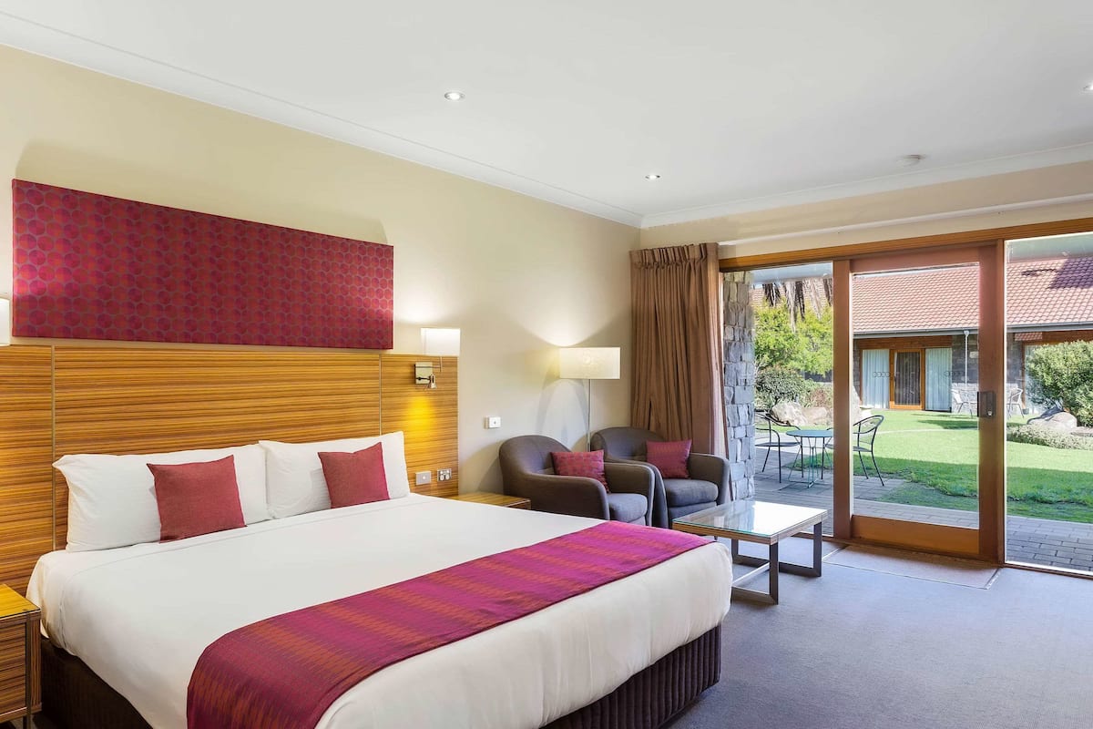 A hotel room with a large bed, two armchairs, a small table, and a sliding glass door leading to an outdoor area.
