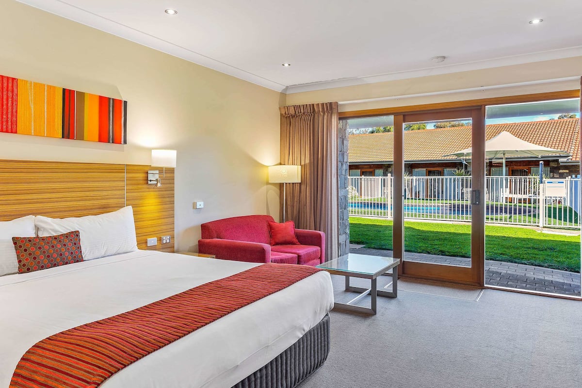 A hotel room with a large bed, a red armchair, a glass coffee table, and a view of a pool and outdoor area.