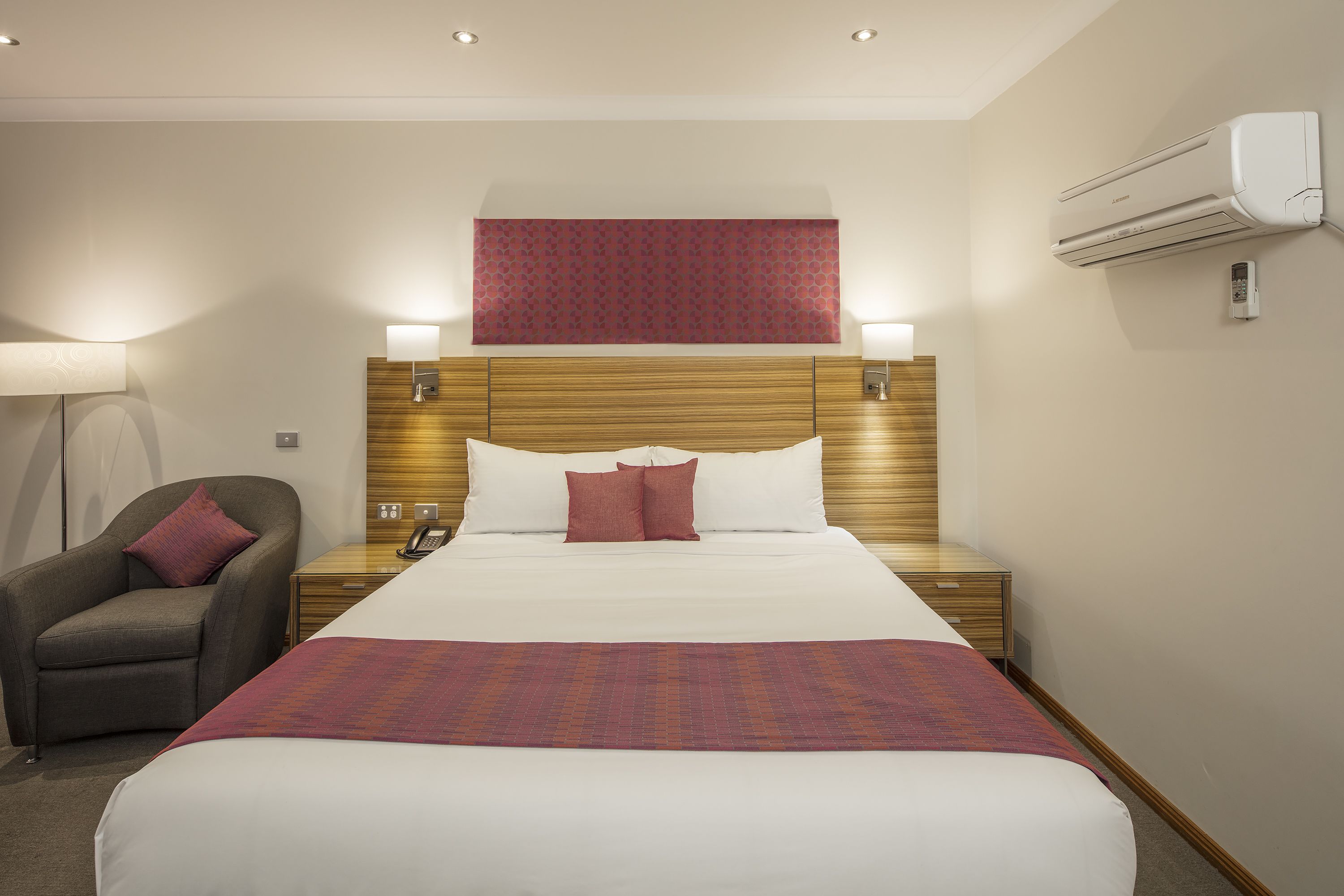 Habitación de hotel con una cama grande, una mesita de noche con lámpara, una silla y un aire acondicionado.