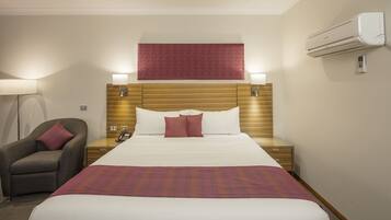 Habitación de hotel con una cama grande, una mesita de noche con lámpara, una silla y un aire acondicionado.