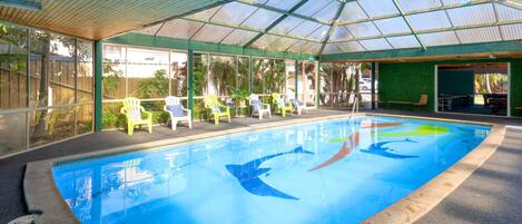 Una piscina interior con agua cristalina y azul, rodeada de sillones de descanso y una zona techada con techo de vidrio.