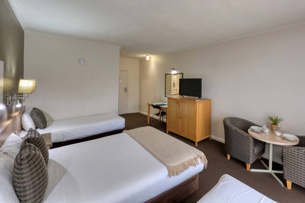 A hotel room with two beds, a desk, a wardrobe, a television, and a small dining table with chairs.