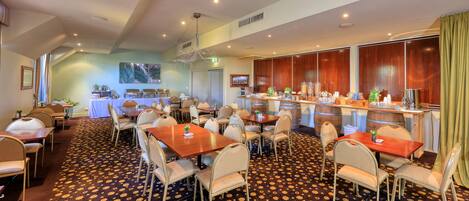 A hotel dining area with multiple tables and chairs, a bar with various food and drink dispensers, and a patterned carpet.