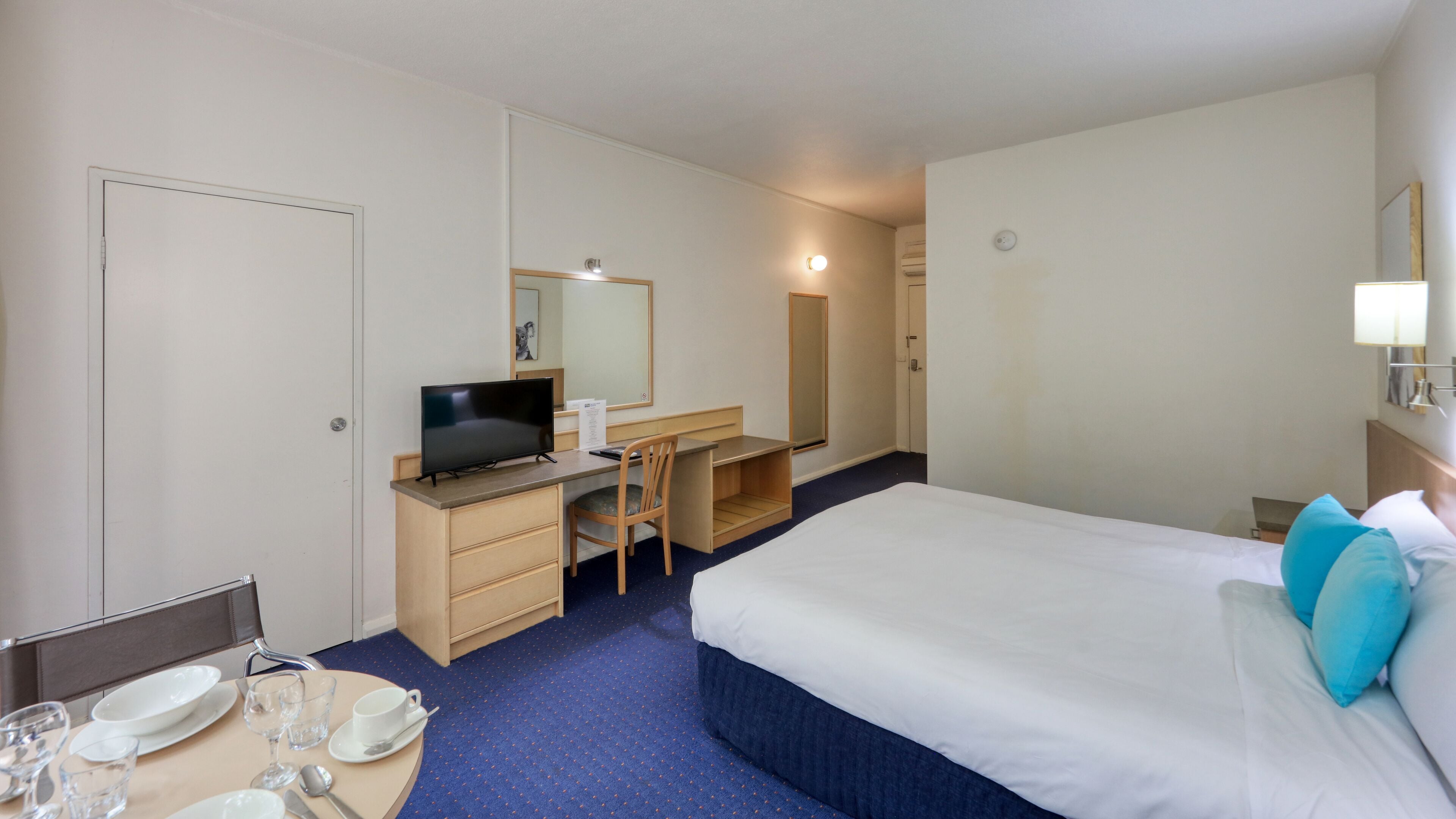 A hotel room with a bed, a desk with a chair, a television, a mirror, and a dining area with a table set for two.