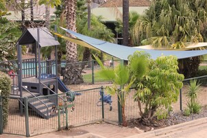 Un parque infantil con tobogán y estructura de escalada, rodeado de palmeras y una zona sombreada.
