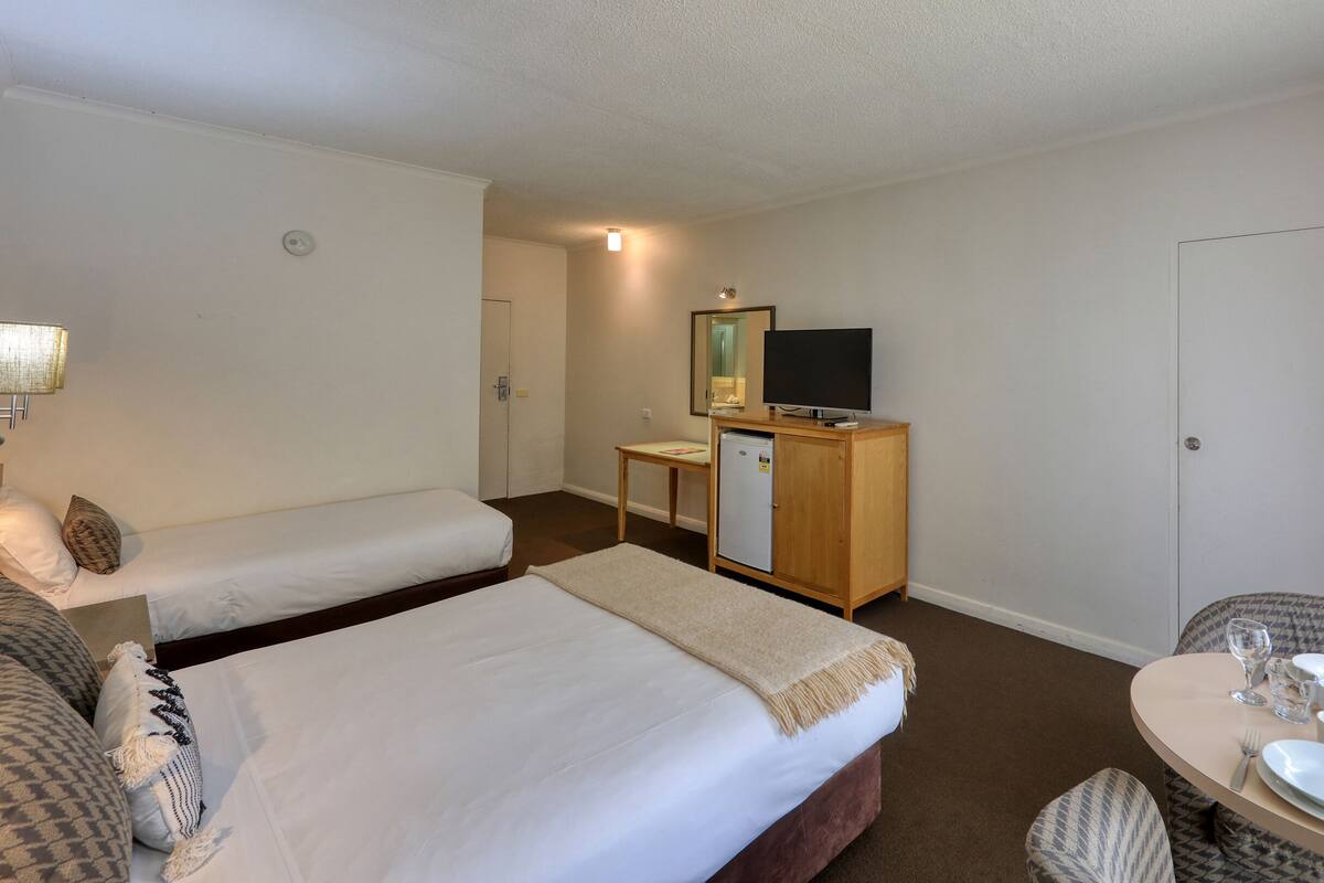A hotel room with two single beds, a small desk with a television, a refrigerator, a dining table set with plates and glasses, and a lamp.
