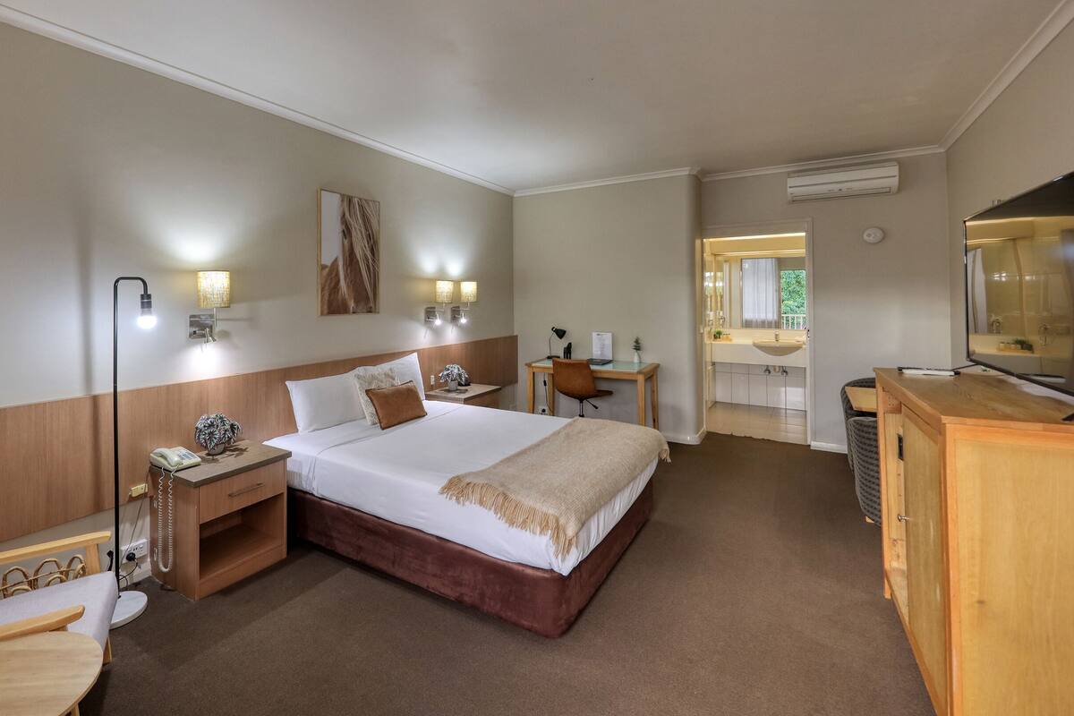 A hotel room with a large bed, bedside table, lamp, chair, desk, and TV.