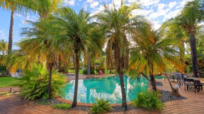 A swimming pool surrounded by palm trees and greenery, with a patio area and outdoor seating. - Swan Hill Resort (Swan Hill)