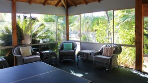 Una habitación con mesa de ping pong, sillas de mimbre y ventanas amplias que dan a vistas de palmeras.