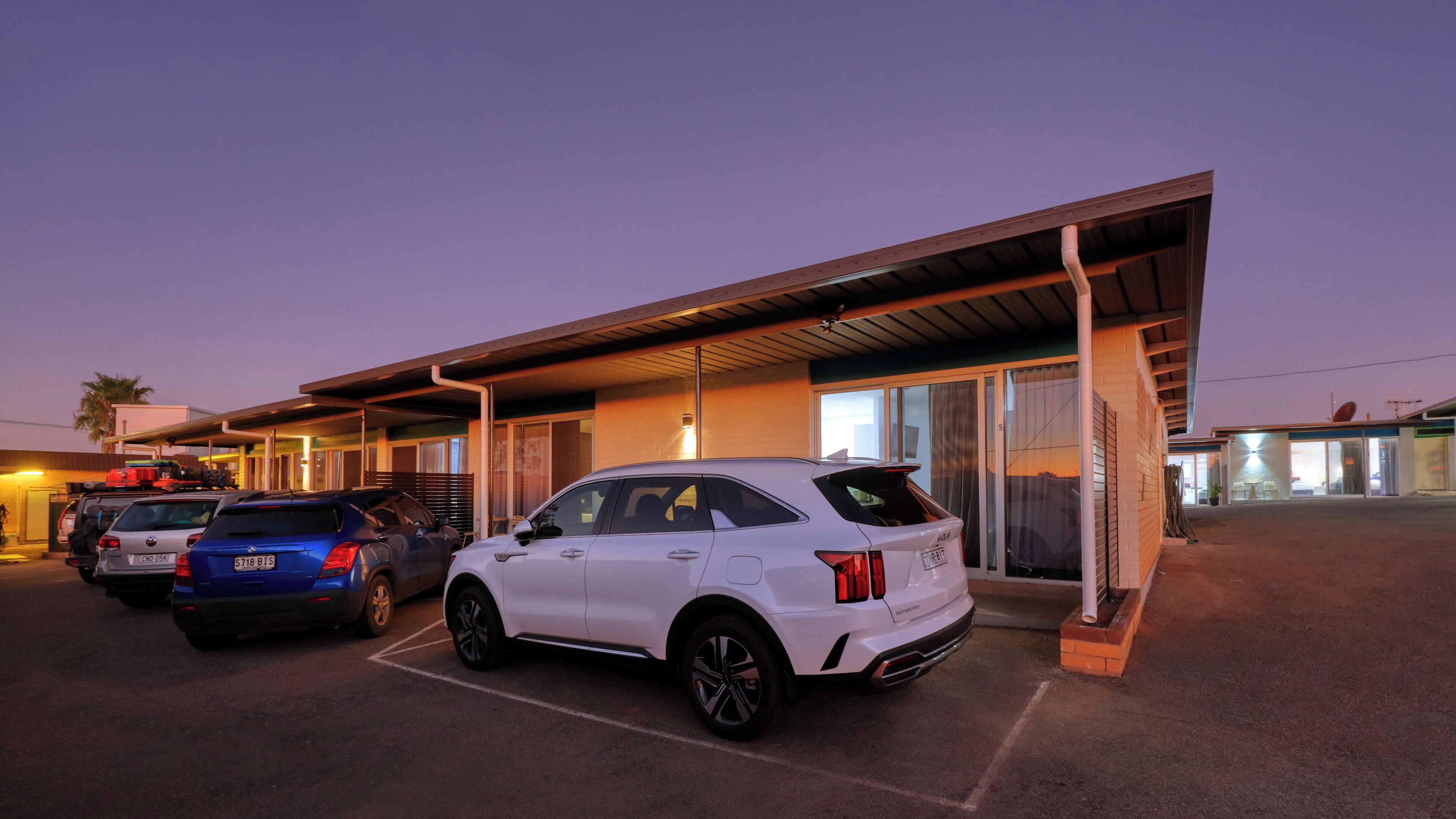 A parking lot with several cars, including a white SUV, parked in front of a building with a covered entrance and large windows.