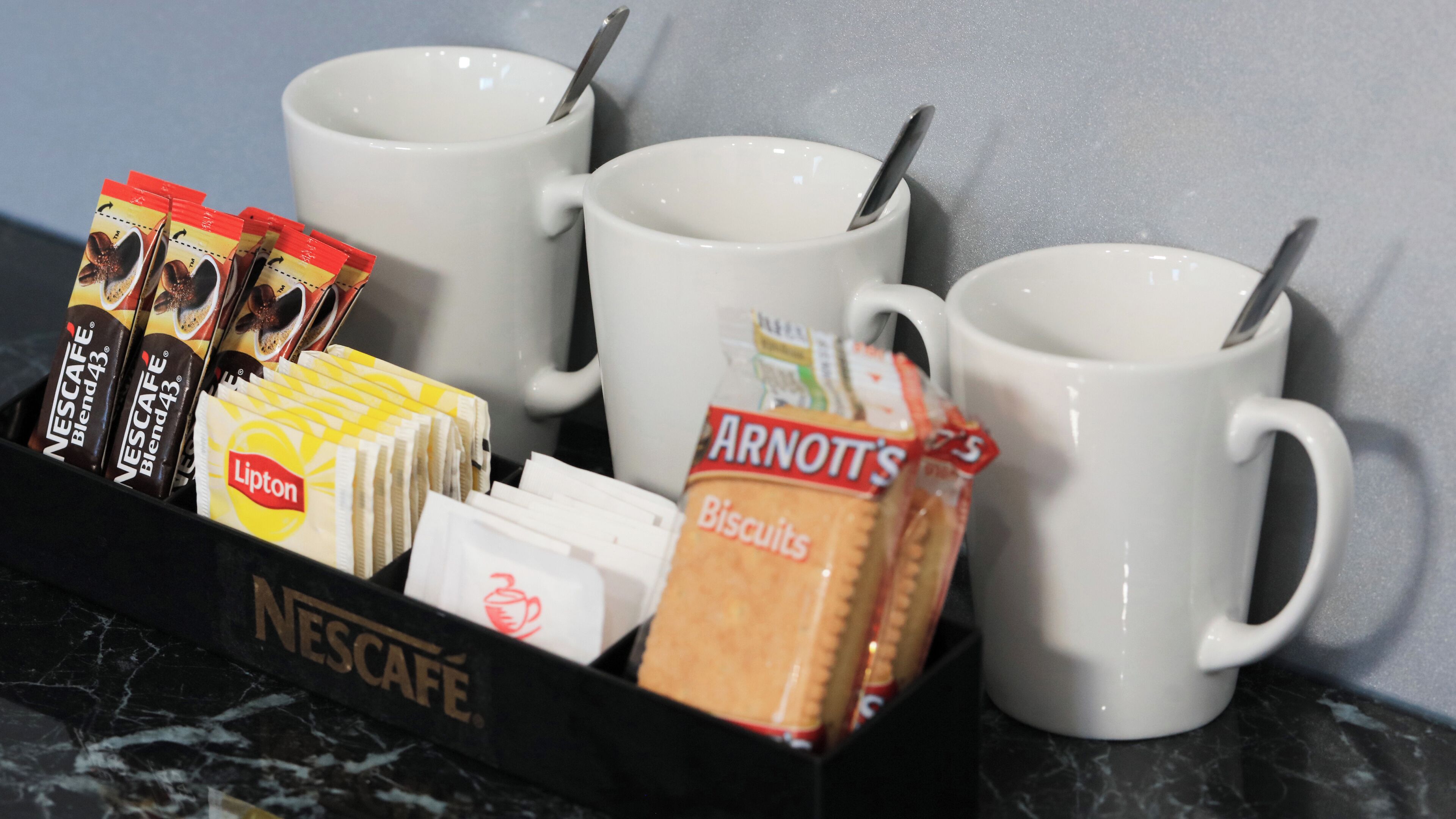 A coffee station with Nescafe Blend 43, Lipton tea, Arnott's biscuits, and two white mugs.