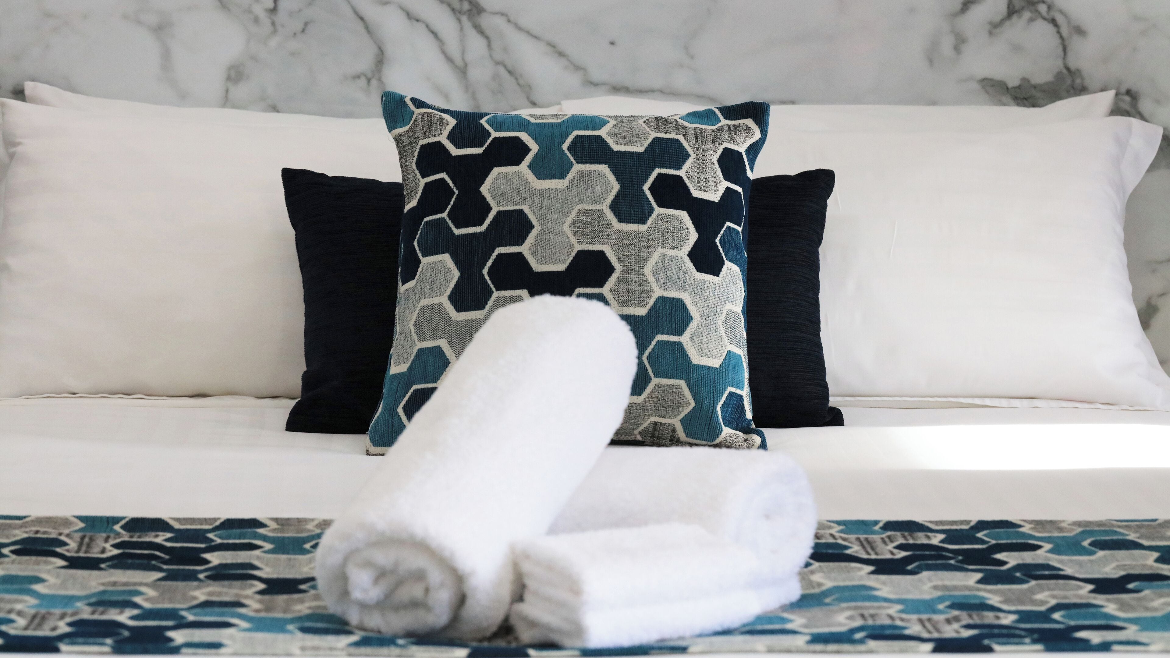 A neatly made bed with a patterned bedspread, two pillows, and a rolled towel on a marble wall background.