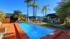 A rectangular swimming pool with a tiled edge, surrounded by a brick border. There are lounge chairs and a table with chairs in the background. - Hilltop Motel (Broken Hill)