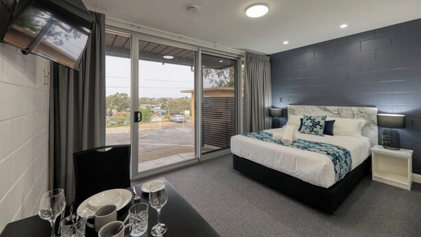 A modern hotel room with a large bed, a dining table set with glasses and plates, and a view of the outside through a sliding door. - Hilltop Motel (Broken Hill)