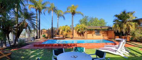 A backyard with a swimming pool, surrounded by palm trees and a brick wall. There is a table and chairs in the foreground.