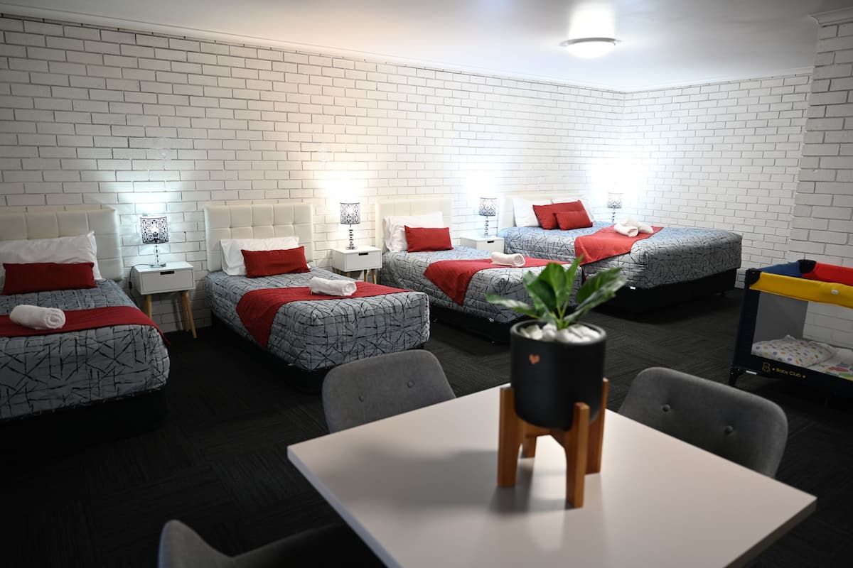 Ett modernt hotellrum med vita tegelväggar, fyra sängar med mönstrat sänglinne, ett matbord och en krukväxt.