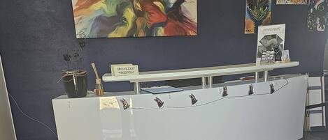 En receptionsdisk med en välkomstmatta med texten ”Välkommen till Camino Inn”, en skylt med texten ”Frukost – menyer” och konstverk på väggen.