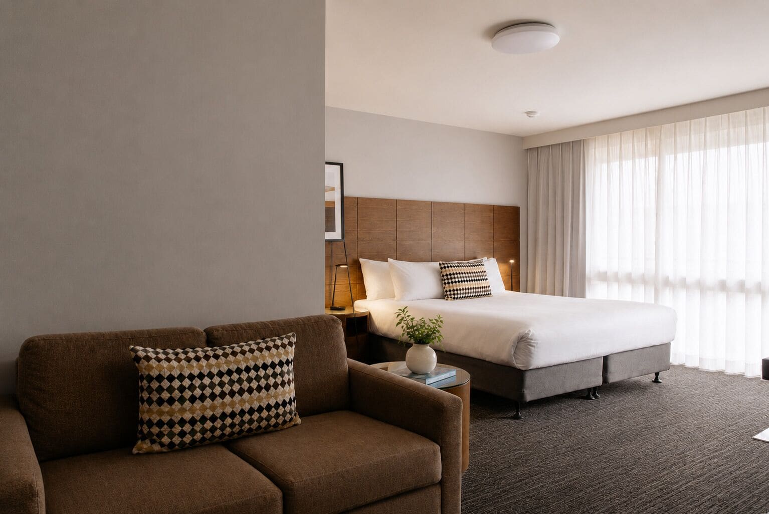 A hotel room with a sofa, a bed with a headboard, a bedside table with a lamp, and a window with curtains.