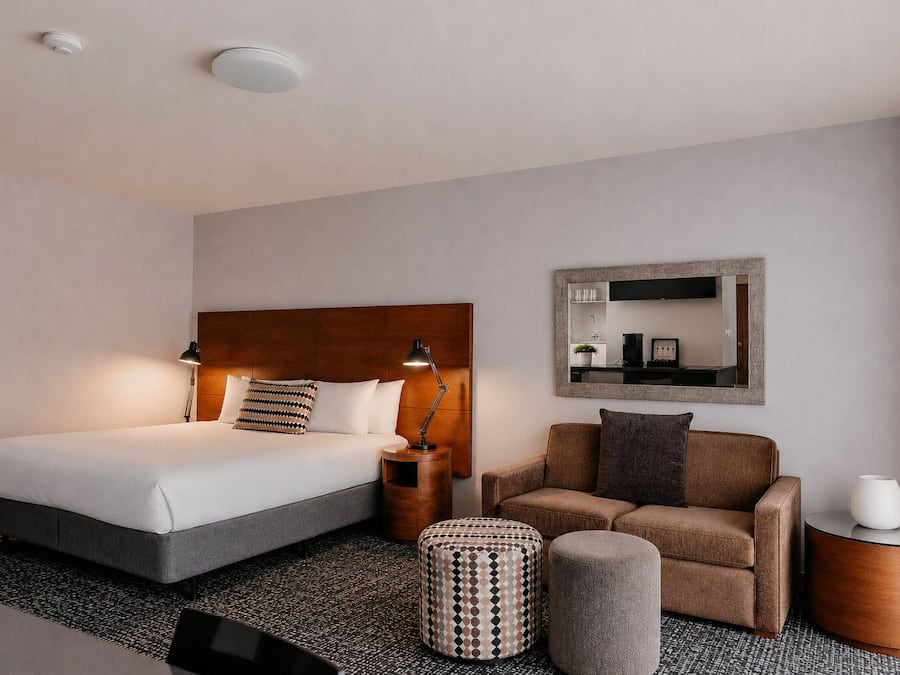 Ein Hotelzimmer mit einem Bett, einem Sofa, einem kleinen Hocker, einem Nachttisch, einer Lampe, einem Spiegel und einer Deckenleuchte.