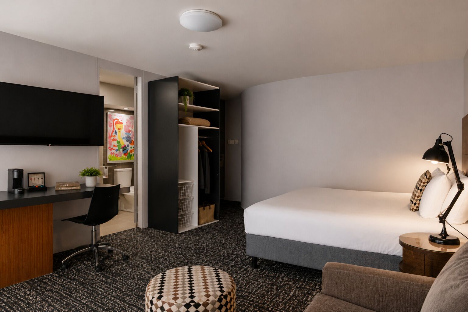 A hotel room with a bed, a desk with a chair, a wardrobe, a television, and a bedside lamp.