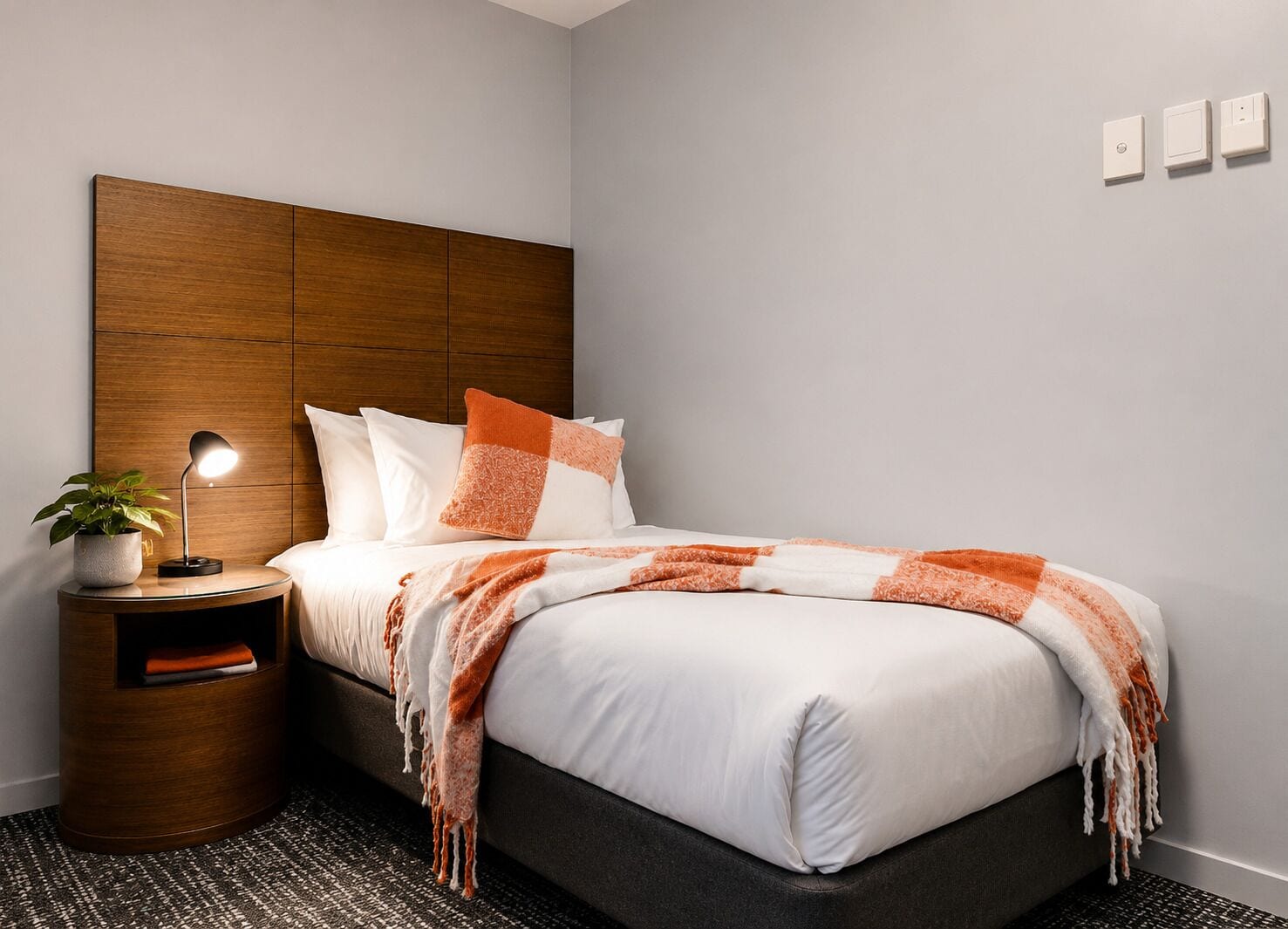 Quarto moderno com cabeceira de madeira, mesa de cabeceira com abajur e cama com roupa de cama branca e detalhes laranja.