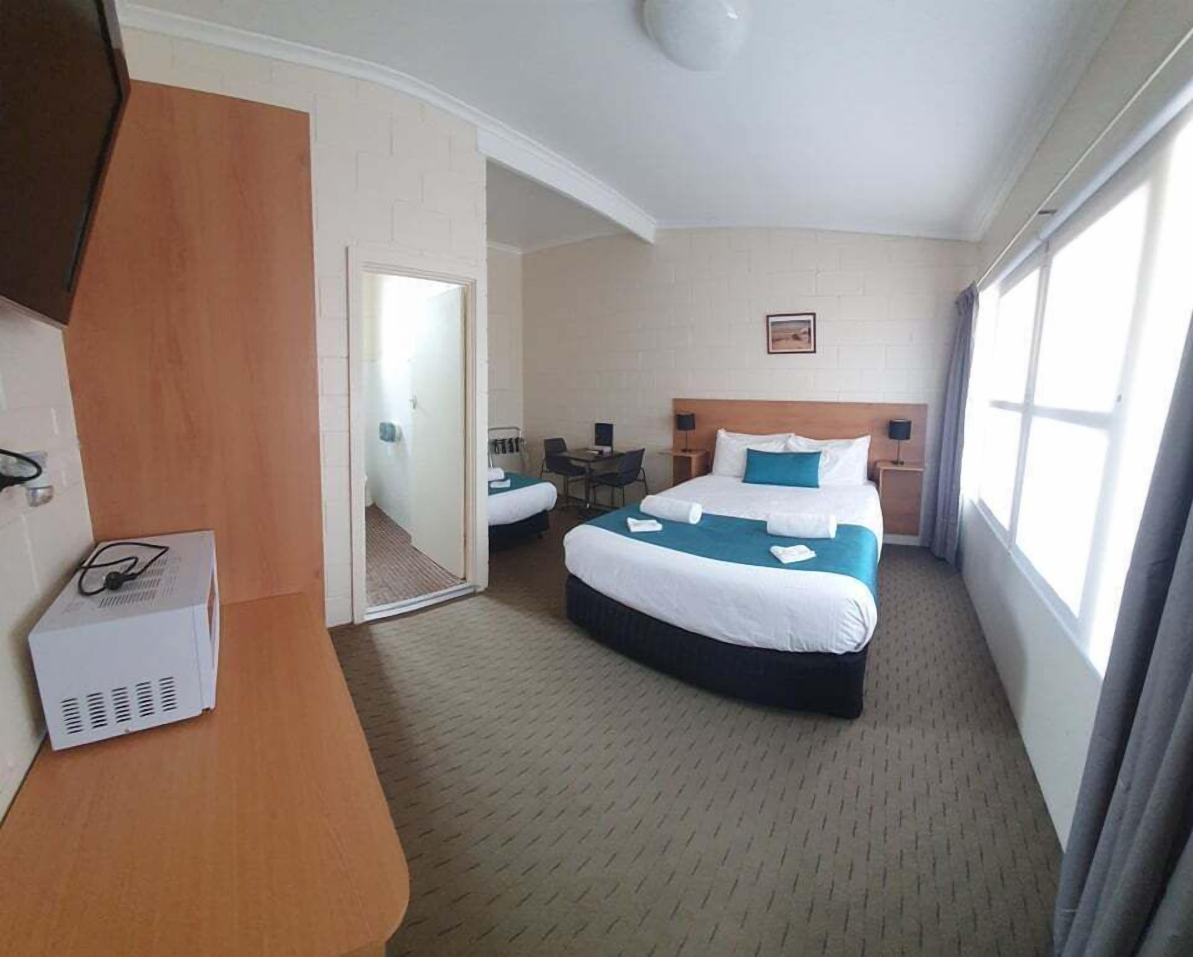 A hotel room with a large bed, a desk, and a chair. There is a mirror on the wall and a window with curtains.