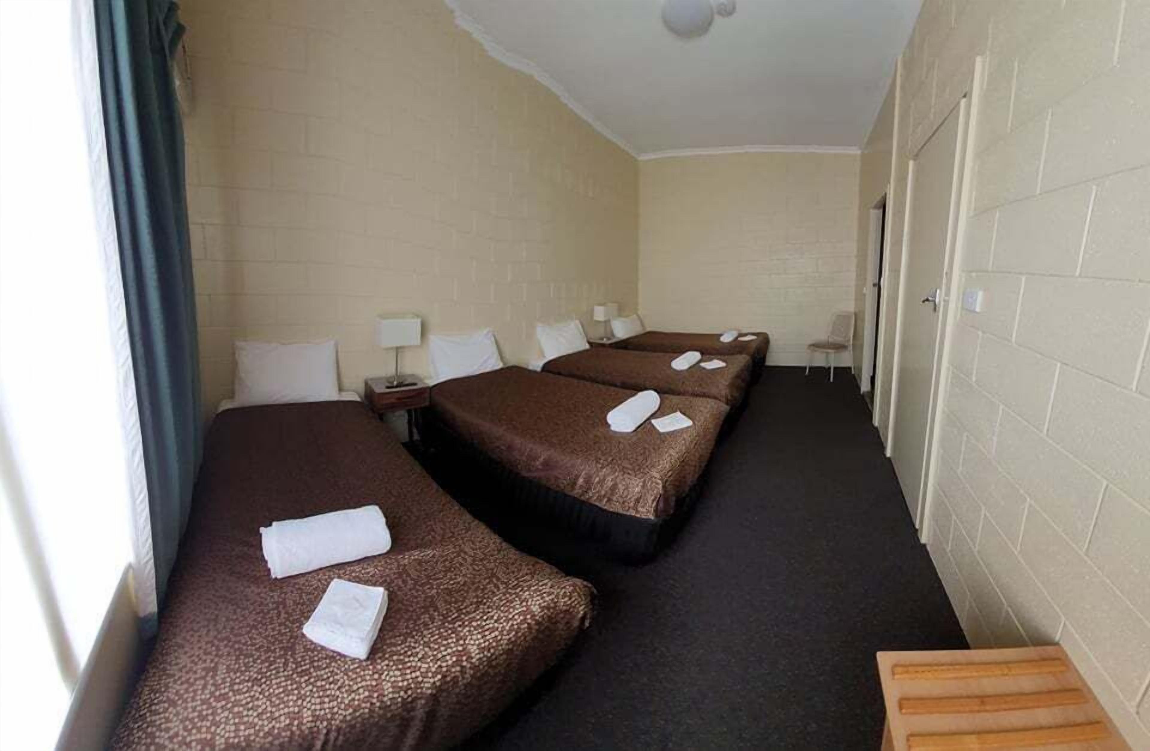 Habitación de hotel con tres camas, cada una con cubrecama marrón y almohadas blancas. Hay una mesita de noche con una lámpara y una silla junto a las camas. El piso está alfombrado y el techo es blanco con una luminaria.